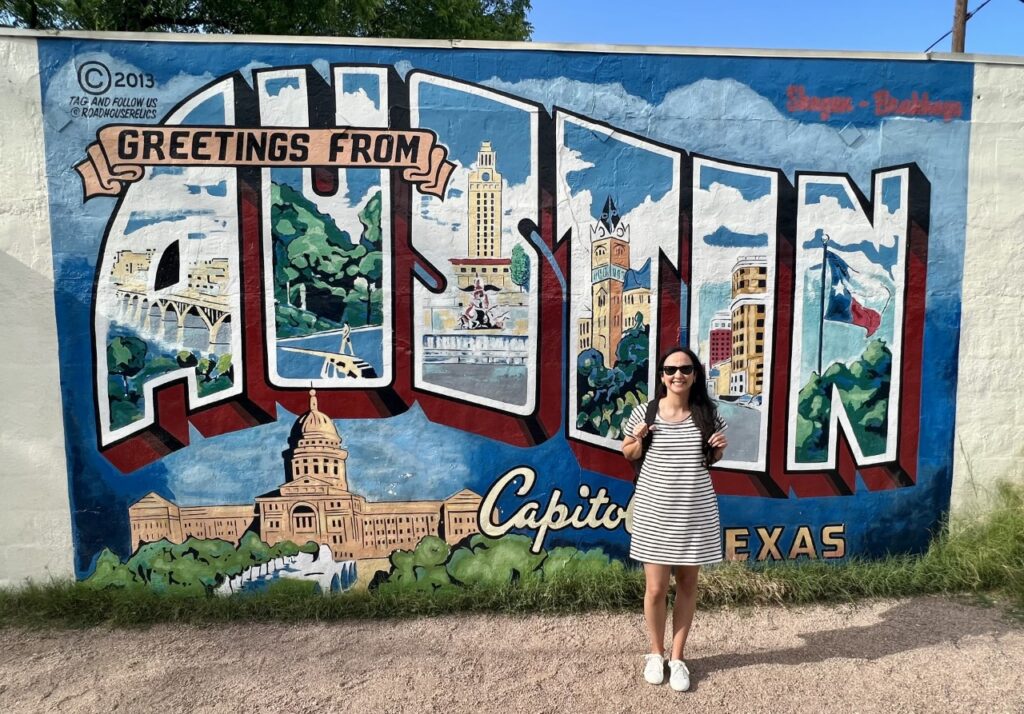 a woman standing in front of the "Greetings from Austin" postcard mural