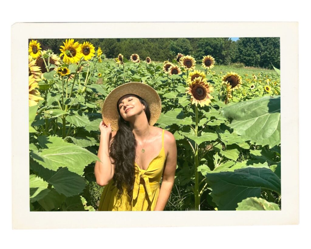 woman standing in a sunflower field holding her hat and smiling with her eyes closed, enjoying the sun on her skin.